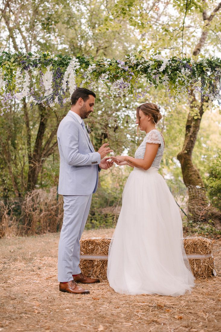 Groom in light blue suit places ring on brides finger under floral arch at rustic outdoor ceremony.