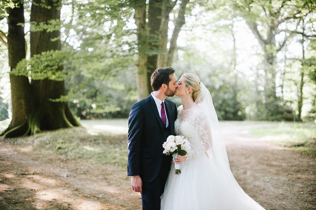 Bride and groom share a tender kiss in a sunlit forest, bride holding a white bouquet.