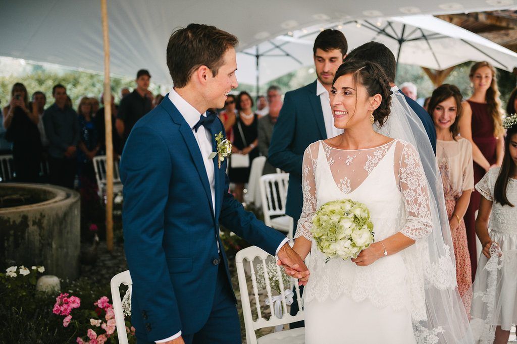 Bride and groom smiling at each other during outdoor wedding ceremony under white tent with guests watching.