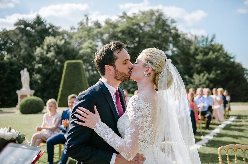 Newlyweds share first kiss as married couple during sunny outdoor garden wedding ceremony.