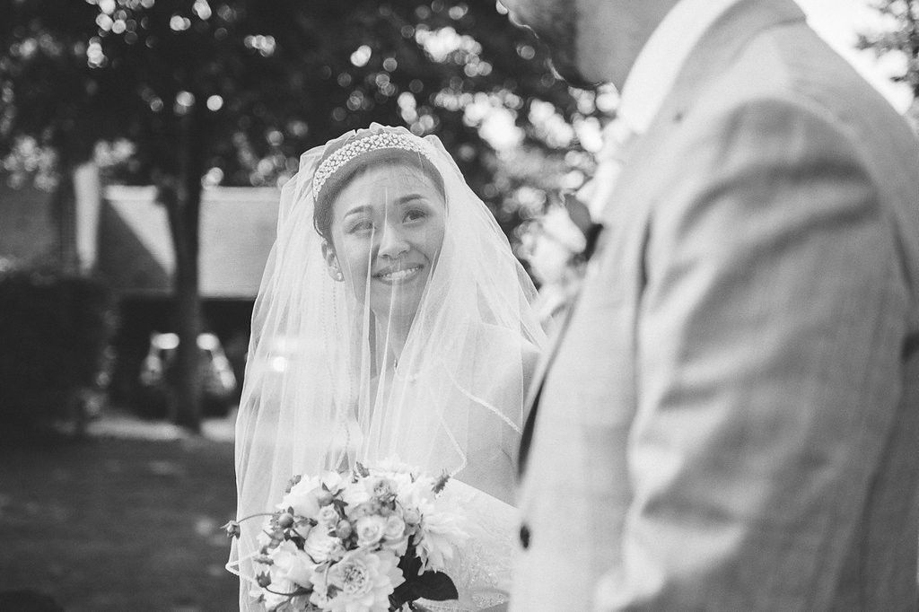 Smiling bride in pearl headband and veil holds white bouquet while gazing at groom outdoors.