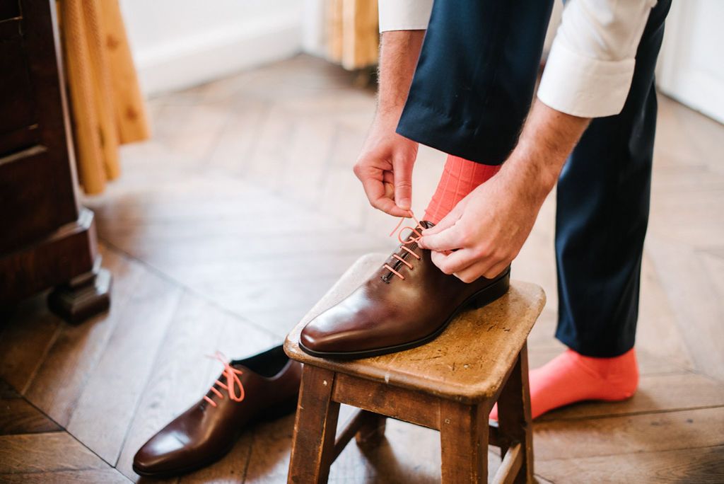 Groom tying brown leather Oxford shoes with coral socks while seated on a wooden stool.