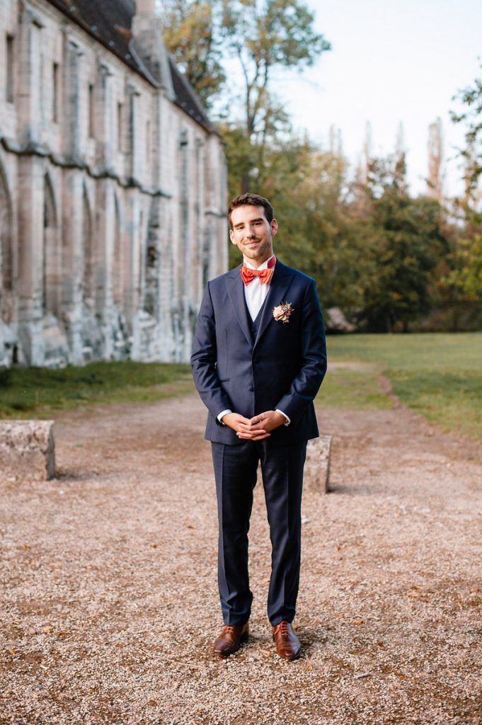 Groom in navy three-piece suit with red bow tie stands smiling outside historic stone building.