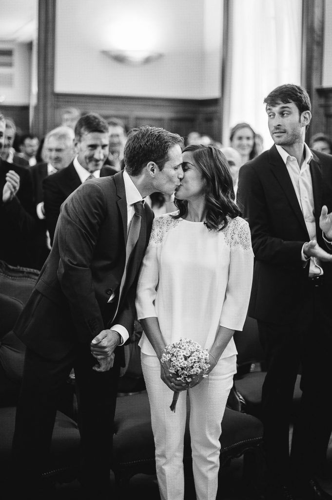 Groom kisses bride in white jumpsuit at wedding ceremony in black and white photo.