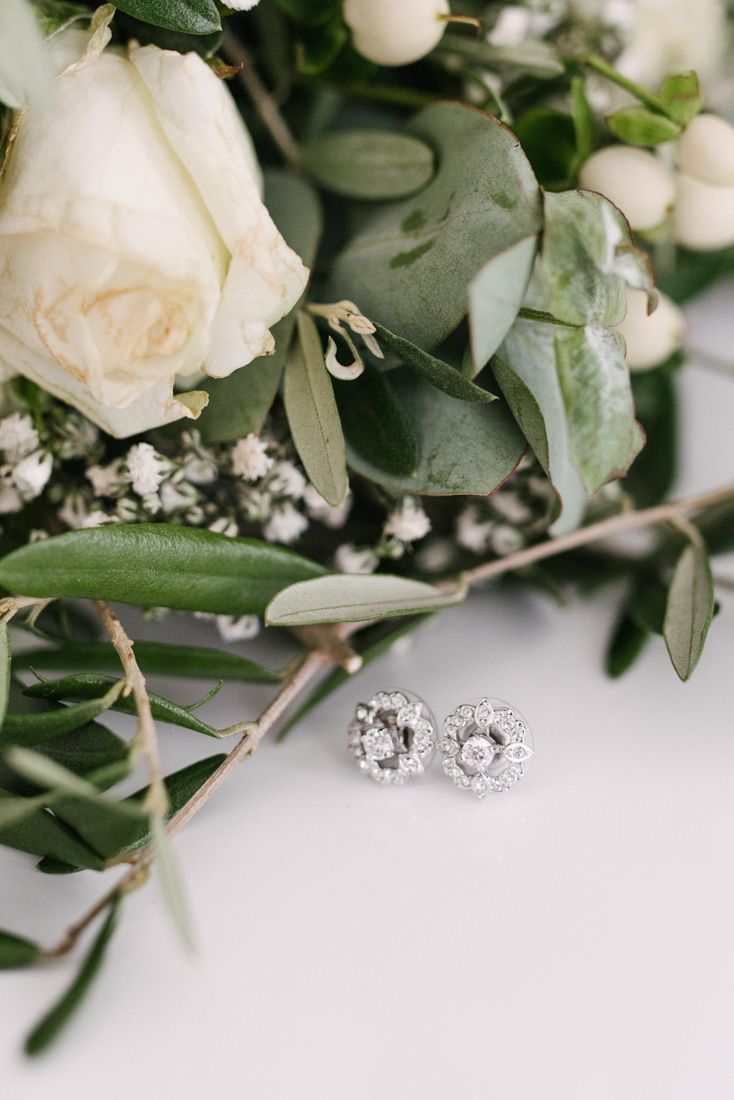 Diamond stud earrings with halo setting displayed beside white roses and greenery wedding bouquet.