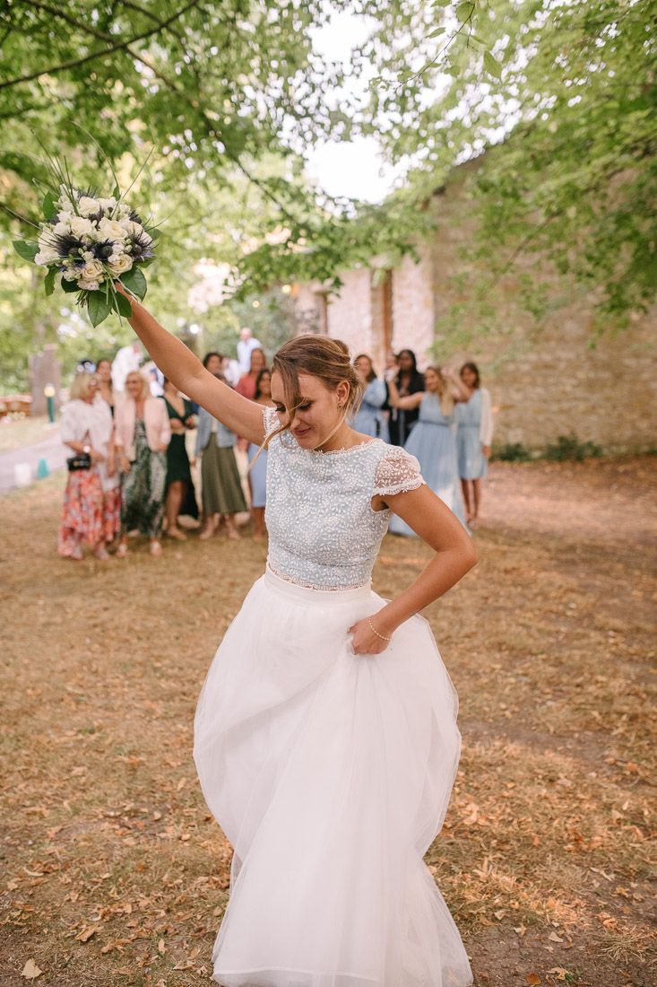 Bride in white tulle gown joyfully tossing bouquet with guests watching in background at outdoor wedding.