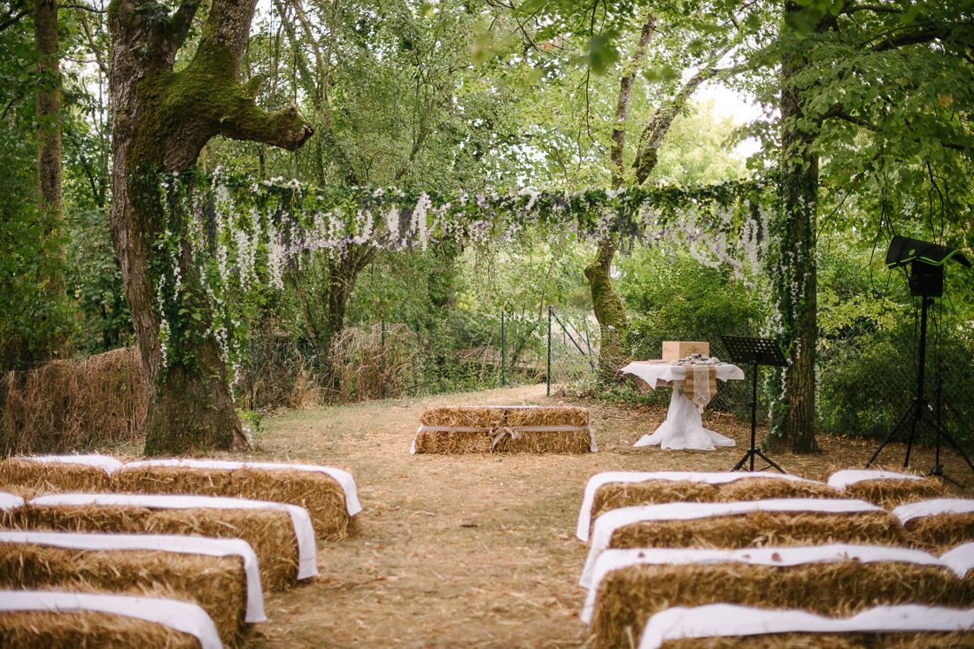 Rustic outdoor wedding ceremony with hay bale seating, white fabric accents, and a draped altar surrounded by lush greenery.