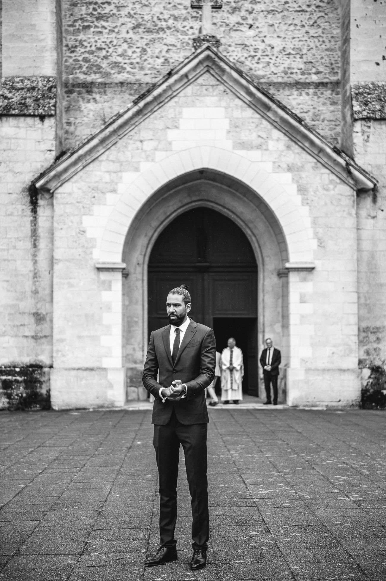 Groom in dark suit stands before stone church arch in black and white wedding photo.