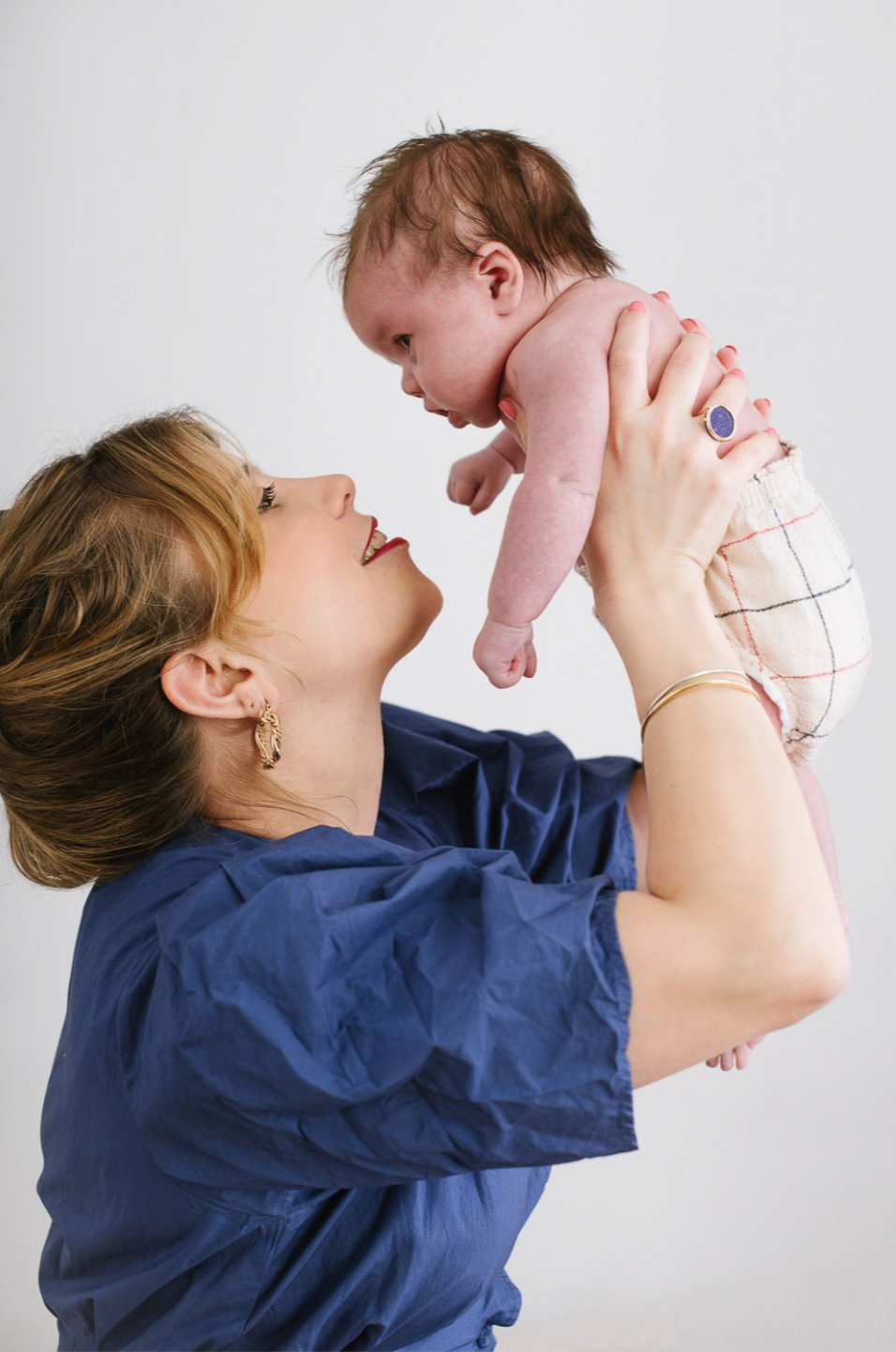 A smiling woman in blue scrubs lifts a newborn baby above her head, gazing up lovingly.