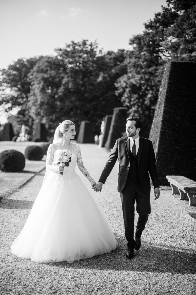 Black and white photo of bride and groom walking hand in hand through elegant formal garden grounds.