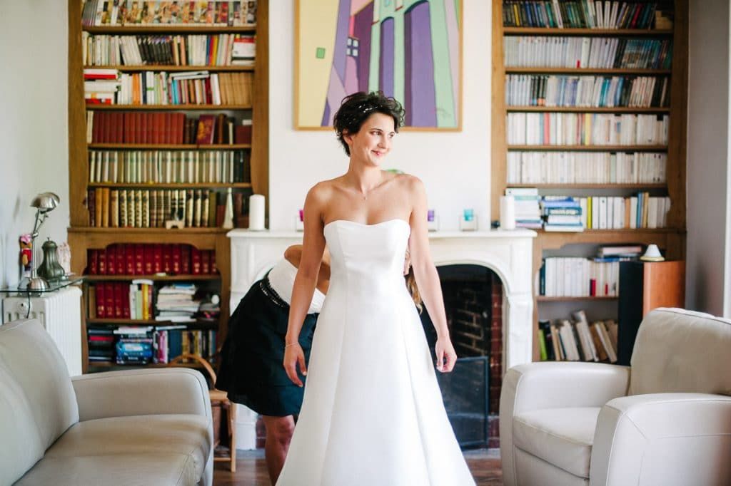 A smiling bride in a strapless white gown stands in a room with bookshelves while someone helps with her dress.