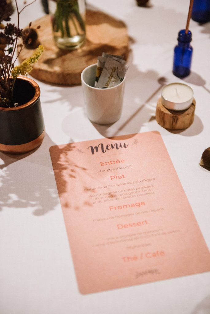 Wedding reception menu card in peach with French text beside small vases and a candle on a white table.