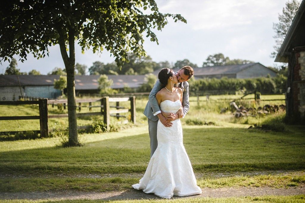 Bride and groom share intimate kiss in golden sunlight near rustic farm fence.