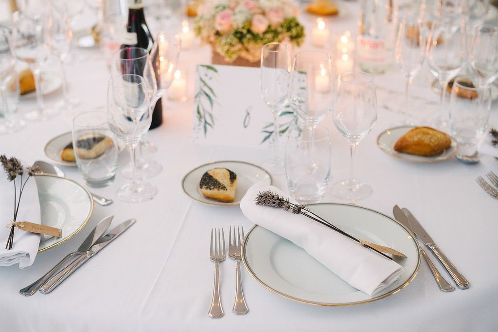 Elegant wedding reception table setting with white linens, gold-rimmed plates, crystal glasses, and candles.
