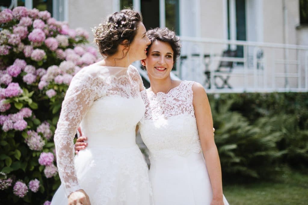 Two brides in white lace wedding dresses share a tender moment in front of pink hydrangea bushes outdoors.