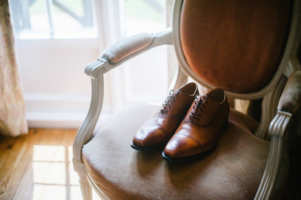 Brown leather oxford dress shoes on vintage upholstered chair by window.
