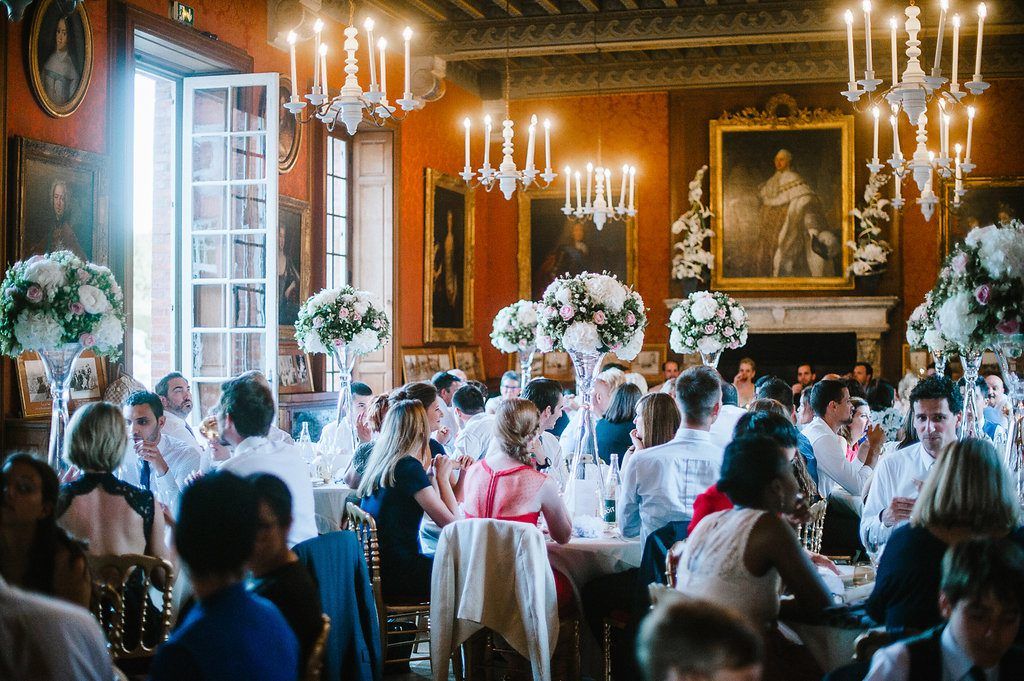 Guests dining at elegantly decorated tables with tall floral centerpieces in grand chandelier-lit chateau.