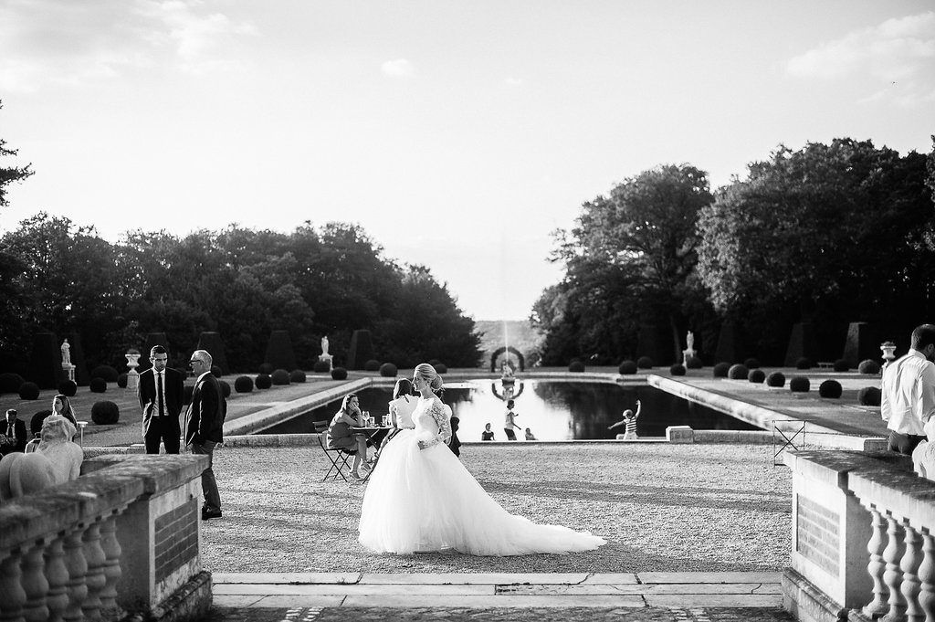 Black and white wide shot of bride in ballgown standing by ornamental reflecting pool at grand estate.