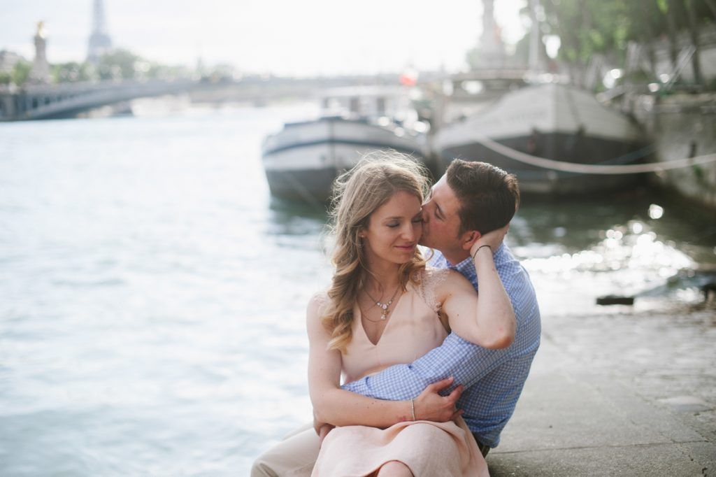 Romantic couple cuddling by the Seine River in Paris with boats and the Eiffel Tower in background.