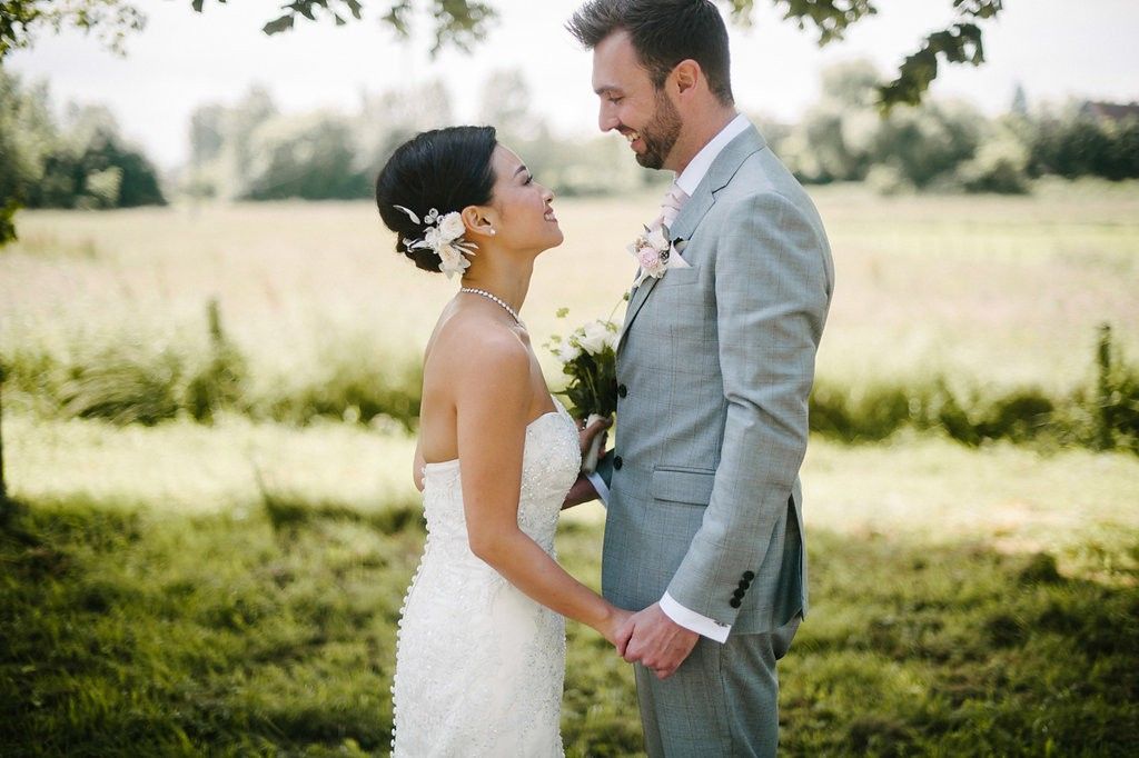 A bride in a white strapless gown and groom in a grey suit hold hands smiling in a sunny green field.