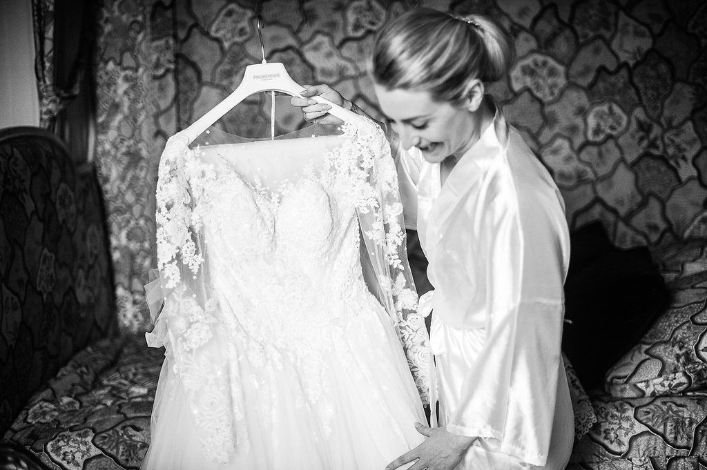 Black and white photo of bride in white robe admiring her lace wedding dress on hanger.