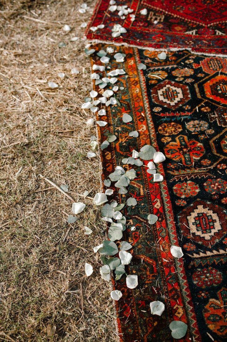 Decorative red Persian rug with scattered white flower petals and eucalyptus leaves on dry grass.