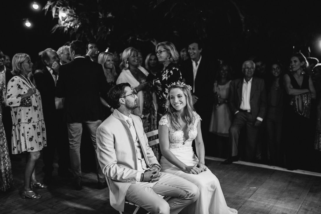 Bride and groom seated together smiling during wedding reception surrounded by guests in black and white photo.