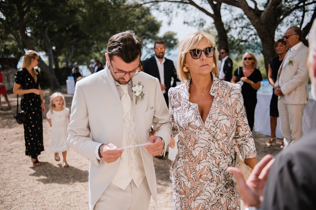 Groom in beige suit reads vows beside elegantly dressed woman in floral dress at outdoor wedding ceremony.
