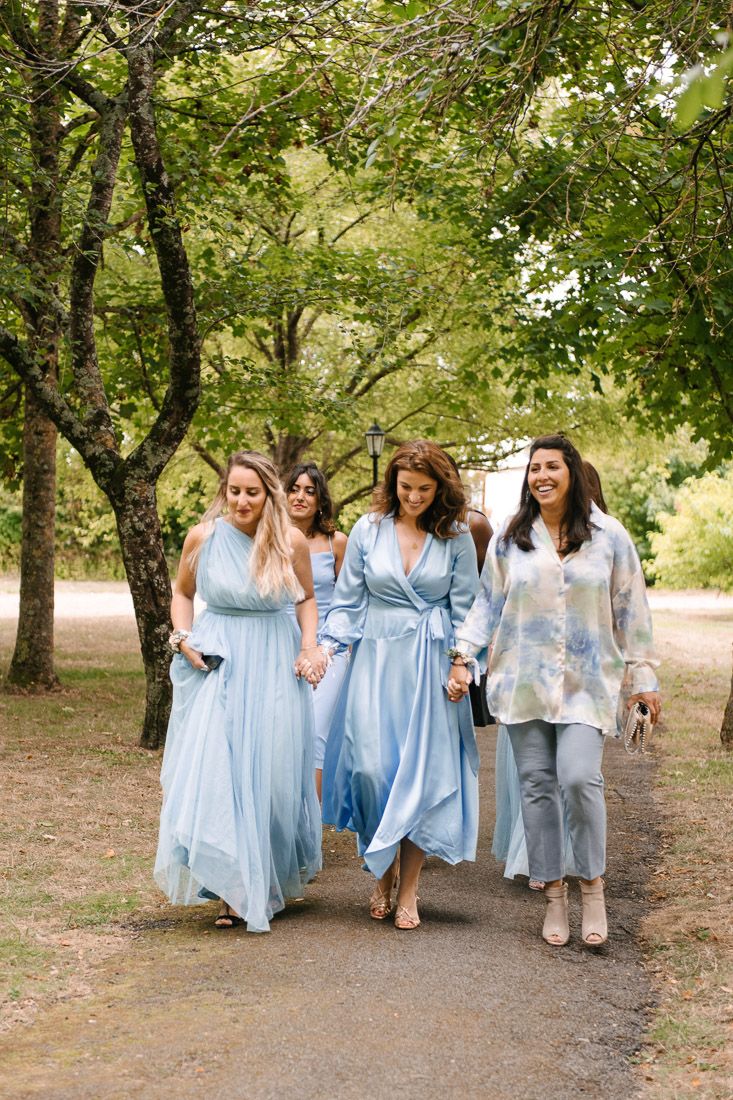 Four women in blue dresses walk together on a garden path surrounded by lush green trees.
