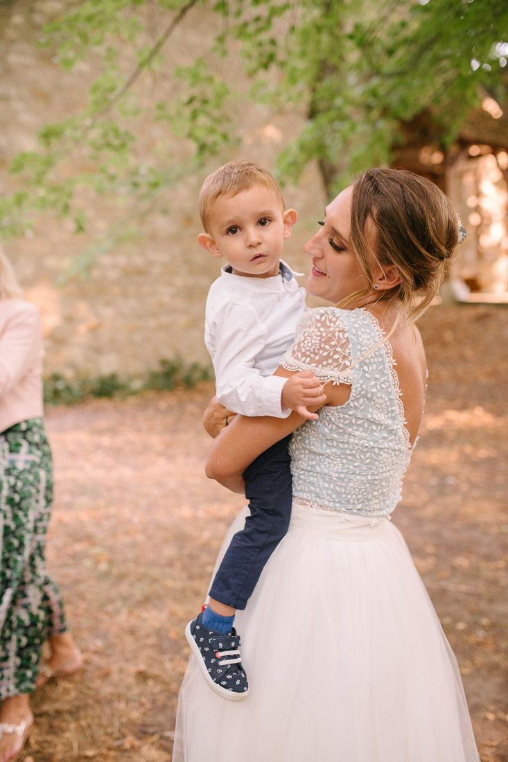 Bride in pearl-embellished wedding gown tenderly holds young toddler boy in white shirt at outdoor wedding reception.