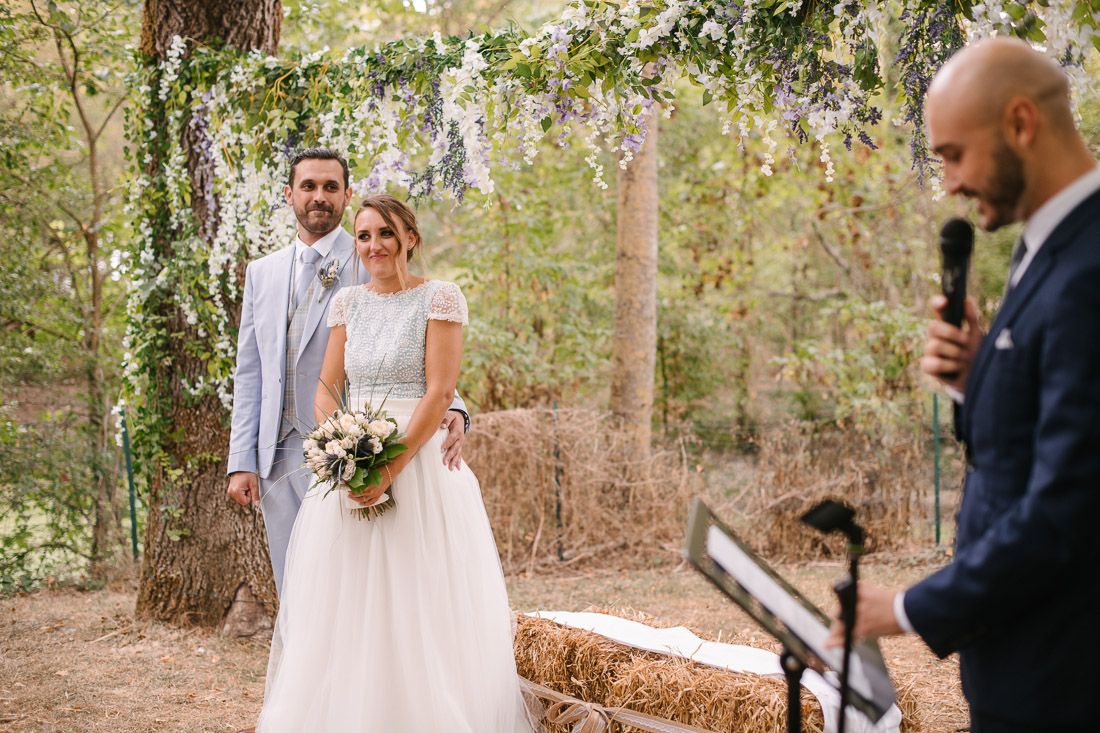 Bride and groom stand together at outdoor wedding ceremony beneath floral arch with officiant speaking at podium.