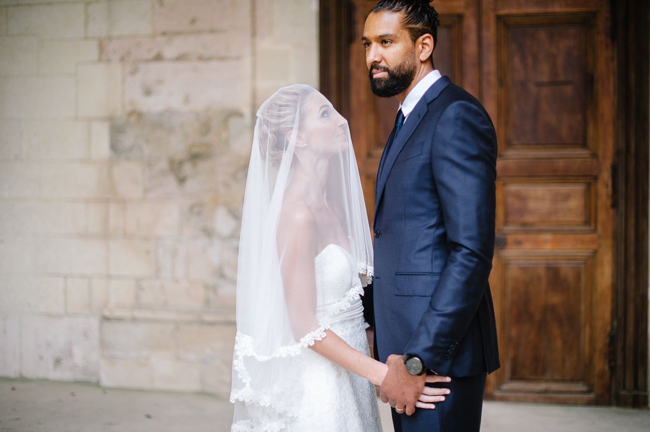Bride in lace veil stands beside groom in navy suit before ornate wooden doors.