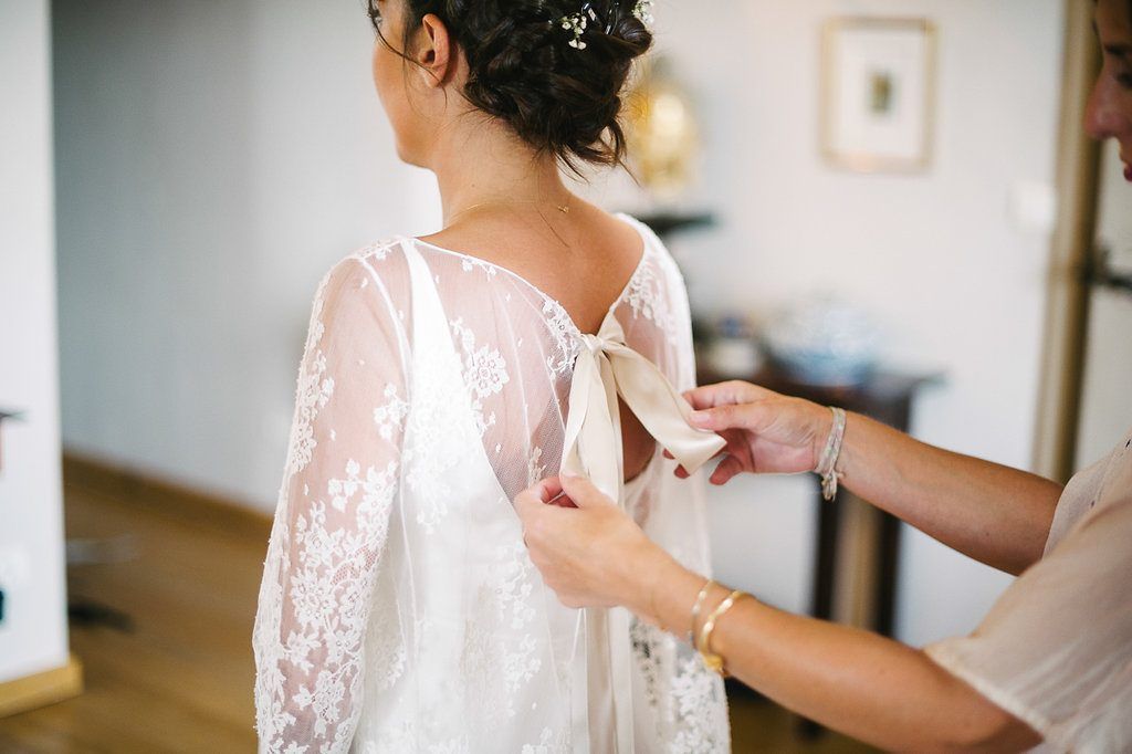 Close-up of bridesmaid tying bow on back of bride's lace long-sleeve wedding dress.