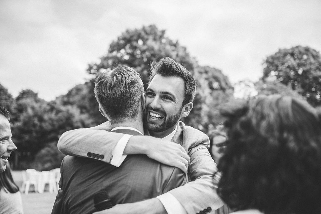 Smiling groom in grey suit shares joyful hug with guest at outdoor wedding reception.