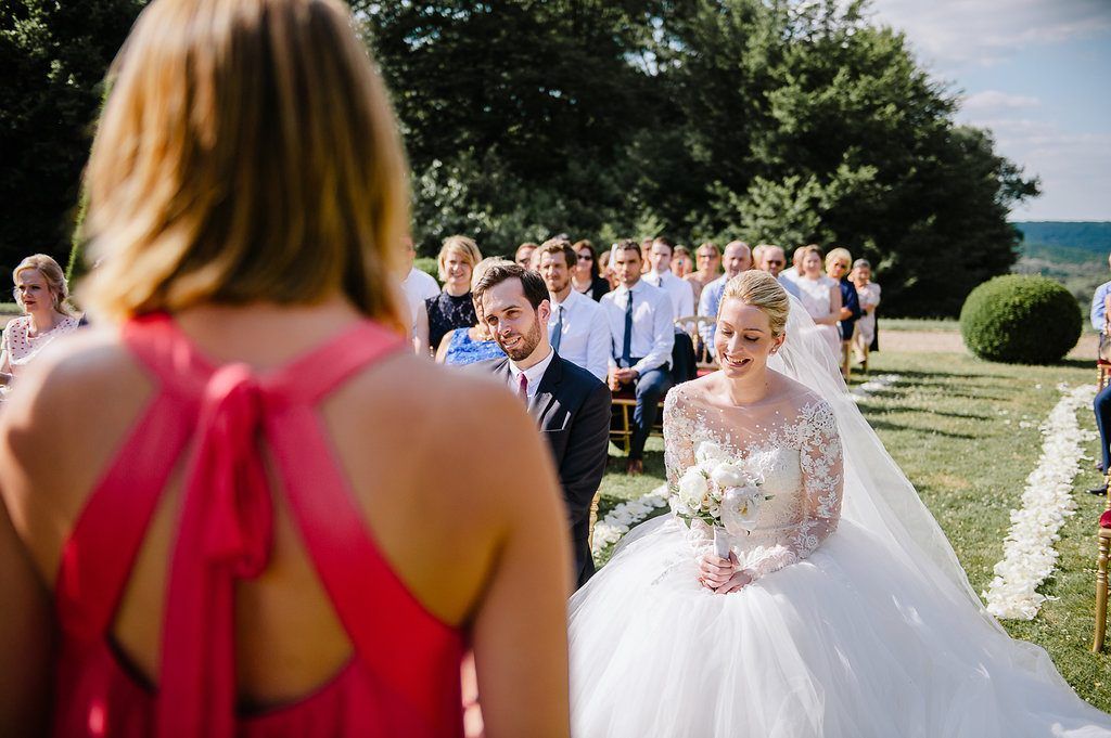 Smiling bride in lace gown watches bridesmaid speak during sunny outdoor garden wedding ceremony.