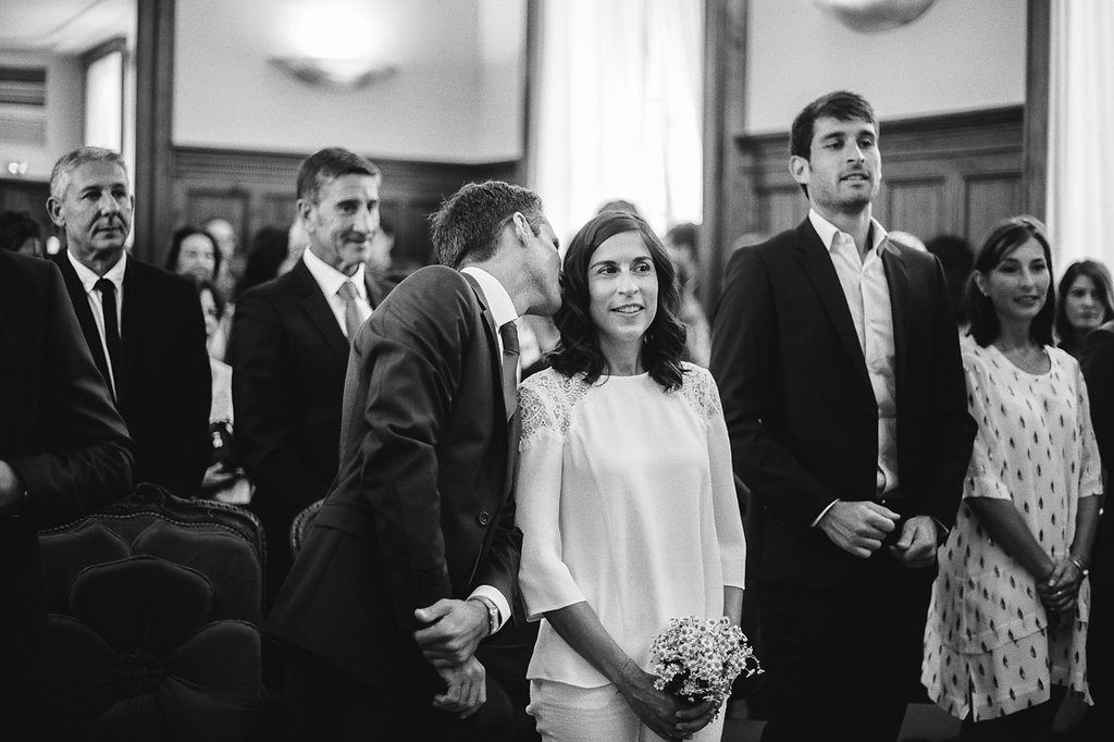 Groom whispers to smiling bride holding bouquet during indoor wedding ceremony black and white.