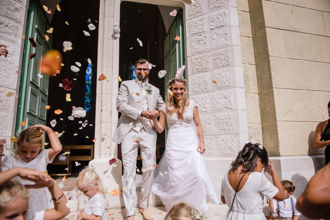 Newlywed couple exits church as guests toss colorful flower petals in joyful wedding celebration.