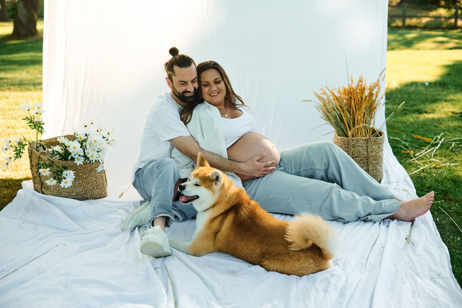 Pregnant couple sitting on white blanket outdoors with Akita dog, surrounded by flowers and wheat decor.