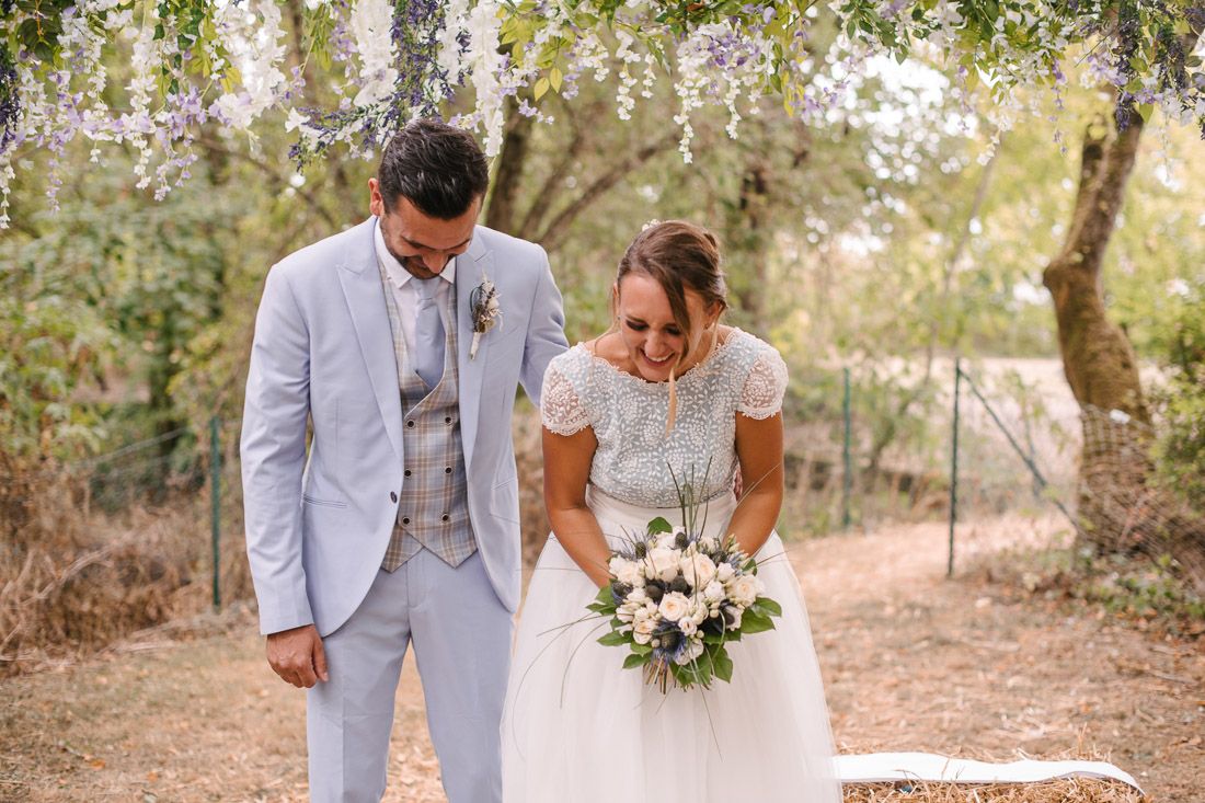 Bride holding bouquet and groom in light blue suit share tender moment at outdoor woodland wedding.