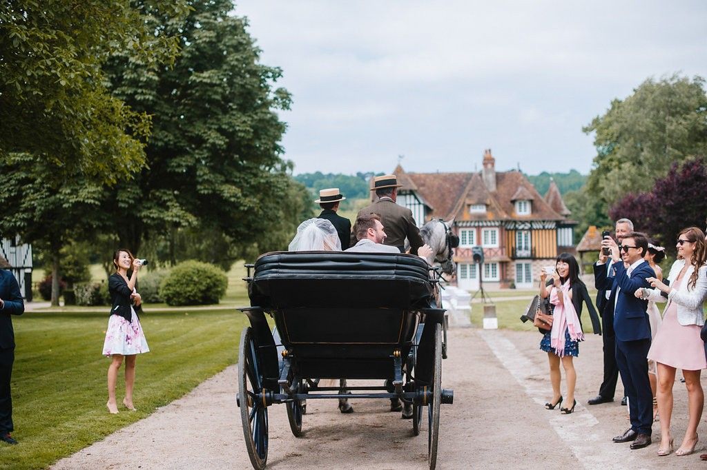 Horse-drawn carriage carries newlyweds past cheering guests on a scenic estate driveway in France.