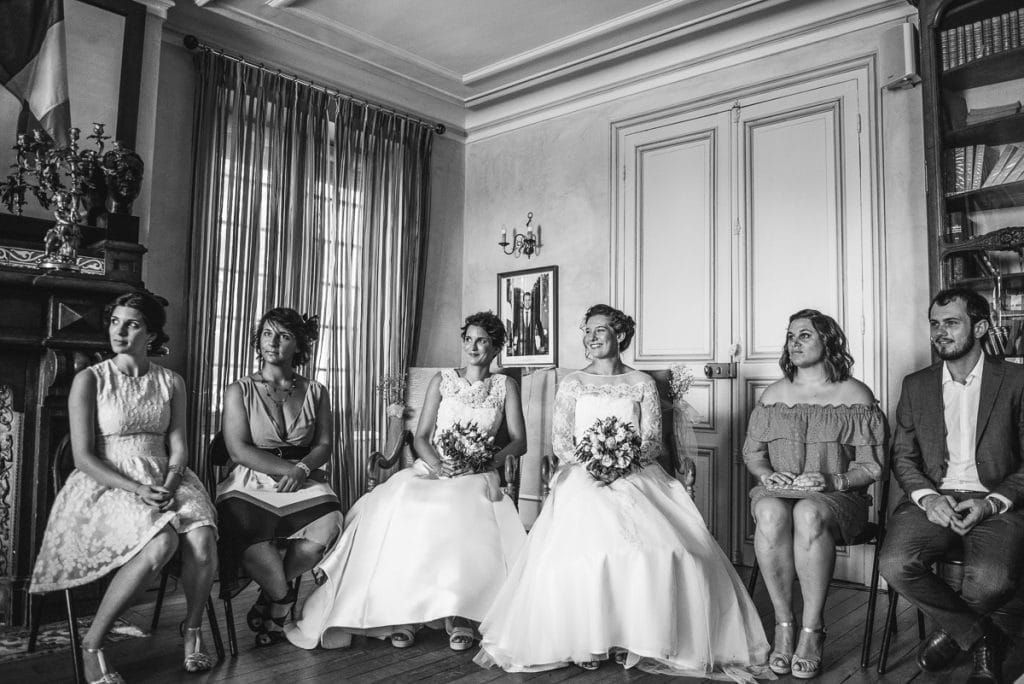 Two brides in white gowns sit with wedding guests in an elegant room during a same-sex wedding ceremony.