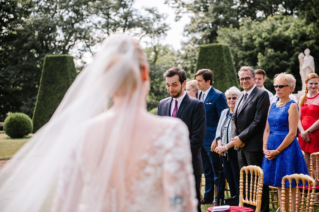 Groom watches emotional bride approach during outdoor garden wedding ceremony with guests seated in golden chairs.
