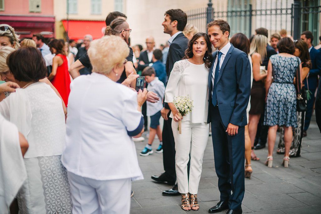 Bride in white jumpsuit and groom in navy suit pose for photo with wedding guests outside.