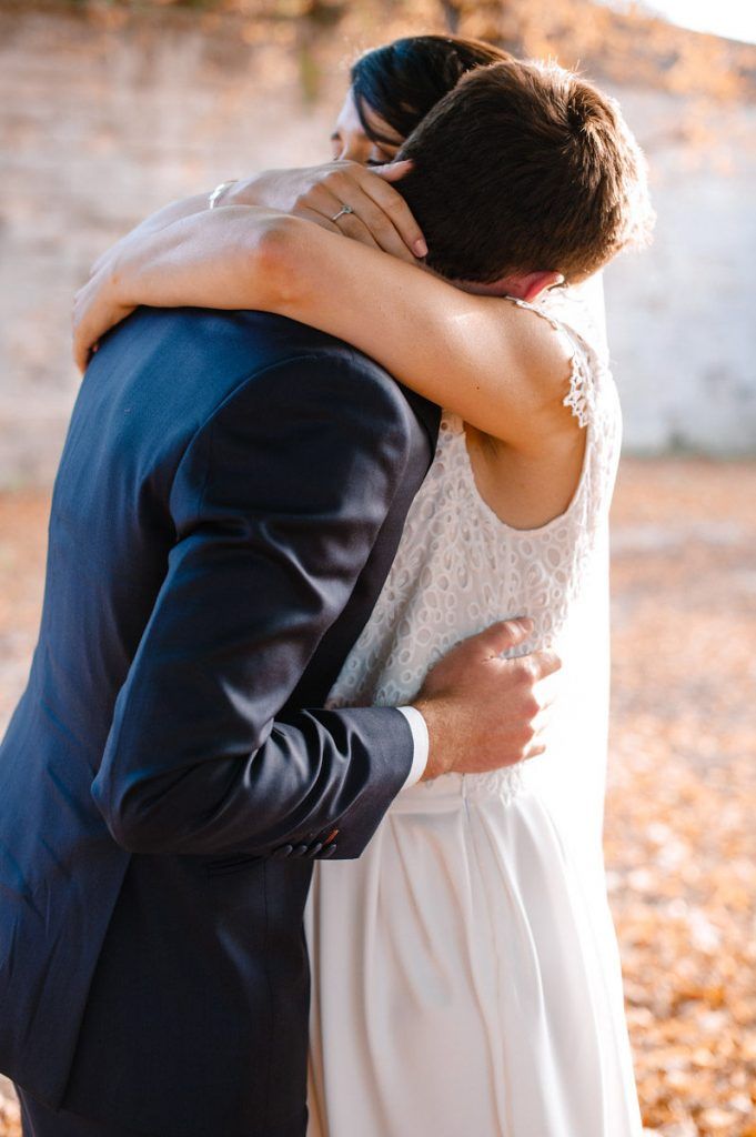 Bride and groom share intimate embrace in warm golden autumn light near stone wall.