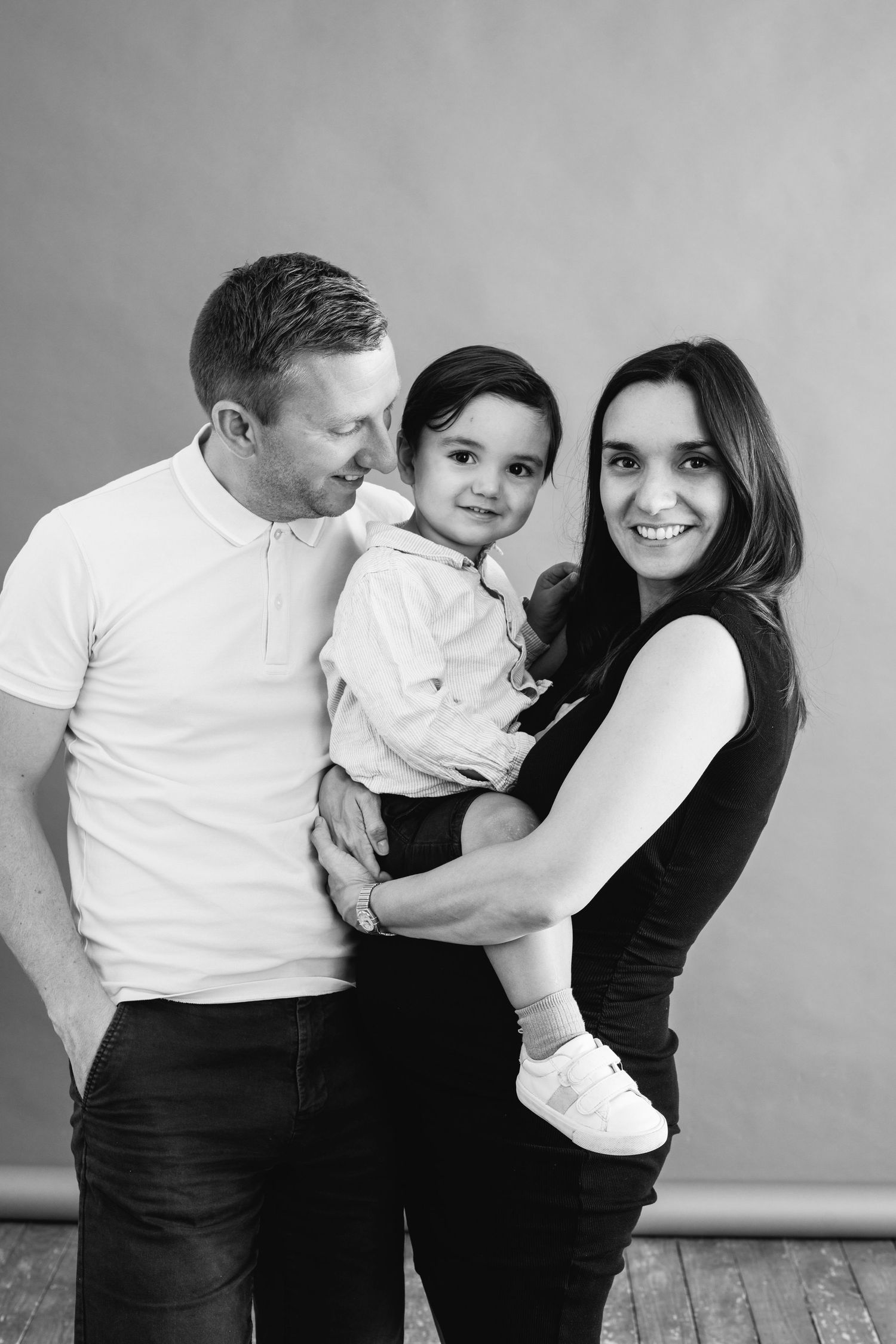 A happy family with parents holding their toddler son in a black and white studio portrait.