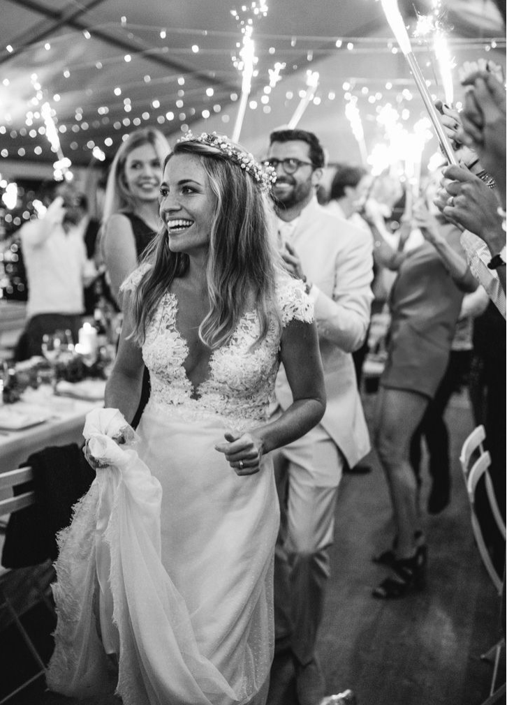 Smiling bride in lace gown and floral crown walks through sparkler tunnel at wedding reception.