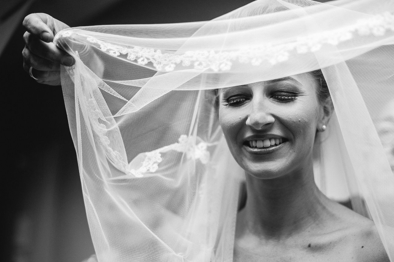 Black and white photo of a smiling bride lifting her lace veil, showcasing elegant bridal beauty.