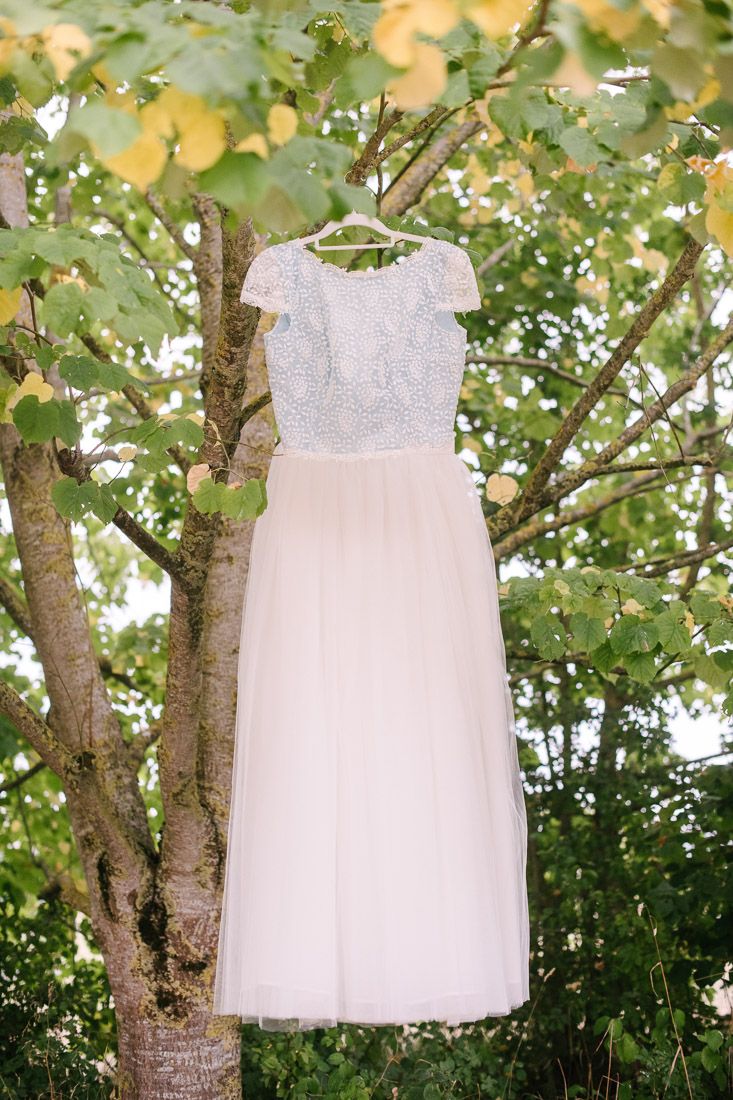 Elegant white lace wedding dress hanging on a hanger from a tree branch outdoors.