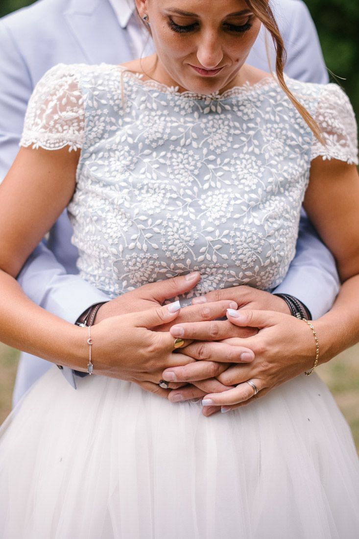 Close-up of bride and groom holding hands together at waist, bride wearing elegant lace wedding gown with gold ring.