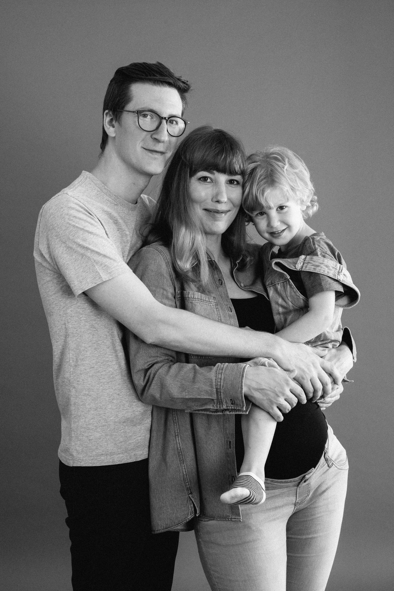 A family of three with parents holding their young child in a black and white studio portrait.